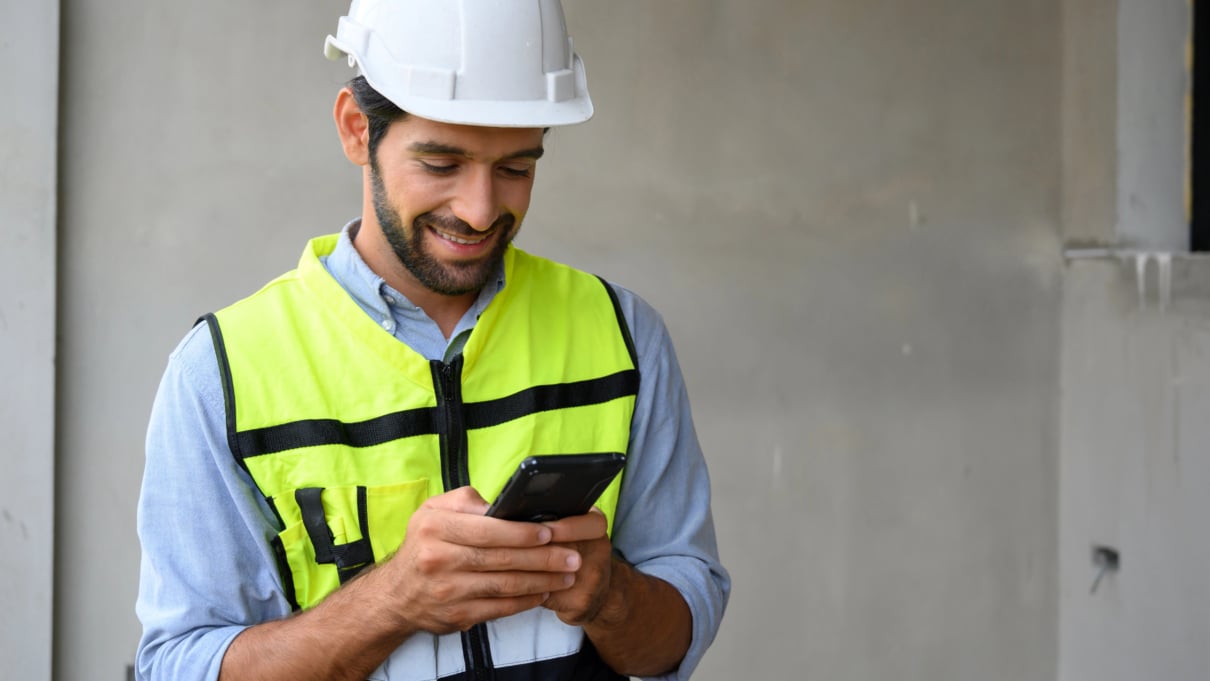 Young man in vest with white helmet standing on construction site using his phone to track assets with Biztags